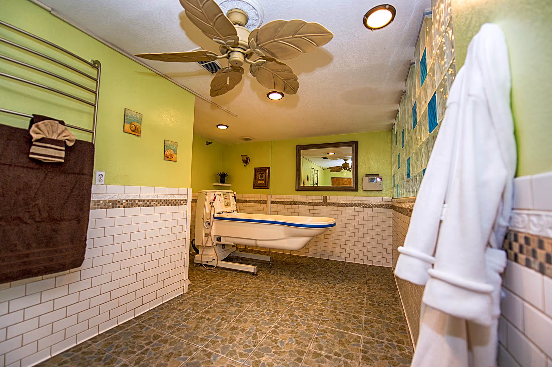 Assisted bathroom with a motorized bathing chair/tub, green walls, tiled floor, towel racks and a ceiling fan.