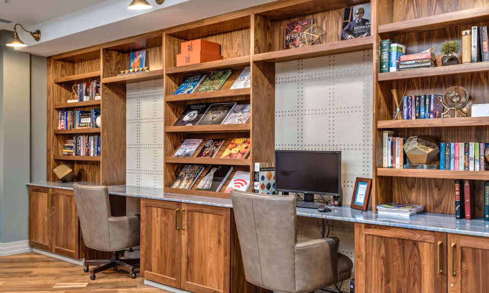 A cozy workspace area with built-in wooden bookshelves filled with books and decorative items. There are two leather swivel chairs in front of a long marble countertop, one with a computer monitor and keyboard. The space is well-lit with wall-mounted lamps and has a warm, inviting atmosphere.