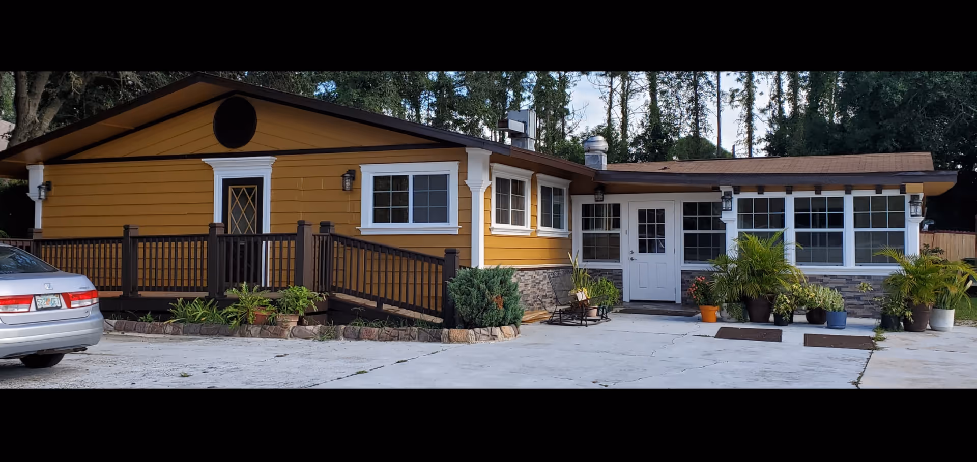 Single-story yellow building front with a ramped wooden porch, white entry door, multiple windows, potted plants and a concrete driveway.