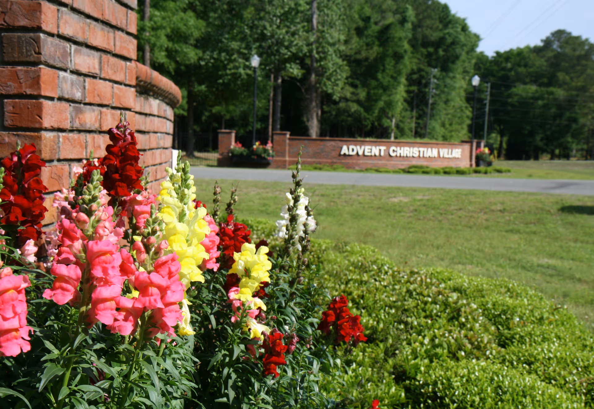 Colorful flowers including pink, red, yellow, and white blooms growing next to a curved brick wall with a grassy area and trees in the background. A brick sign in the distance reads 'ADVENT CHRISTIAN VILLAGE'.