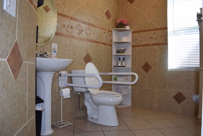 A bathroom with beige tiled walls and floor, featuring a white toilet with safety grab bars on both sides, a white pedestal sink with a round mirror above it, a small white shelving unit with toiletries and towels, and a window with blinds letting in natural light.