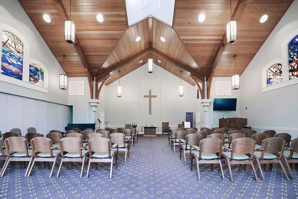 Interior view of a chapel with wooden vaulted ceiling, stained glass windows on both sides, rows of wooden chairs with light cushions arranged facing a wooden altar with a cross mounted on the wall behind it. Pendant lights hang from the ceiling, and there is a television mounted on the right wall.