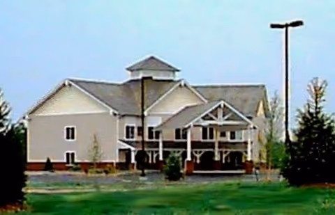 Exterior view of a two-story building with a gabled roof and a covered entrance, surrounded by grass and trees under a clear sky.