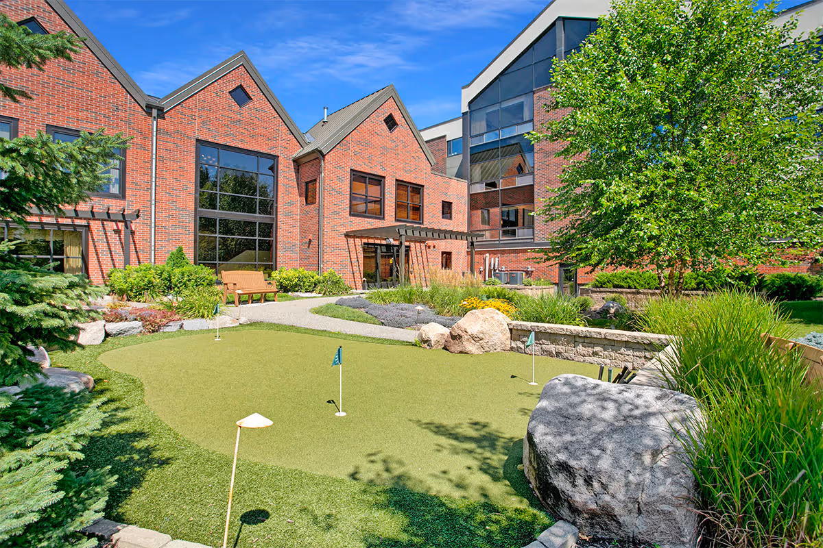 Outdoor putting green area with small flags and holes surrounded by landscaping, rocks, and greenery in front of a red brick building under a clear blue sky.