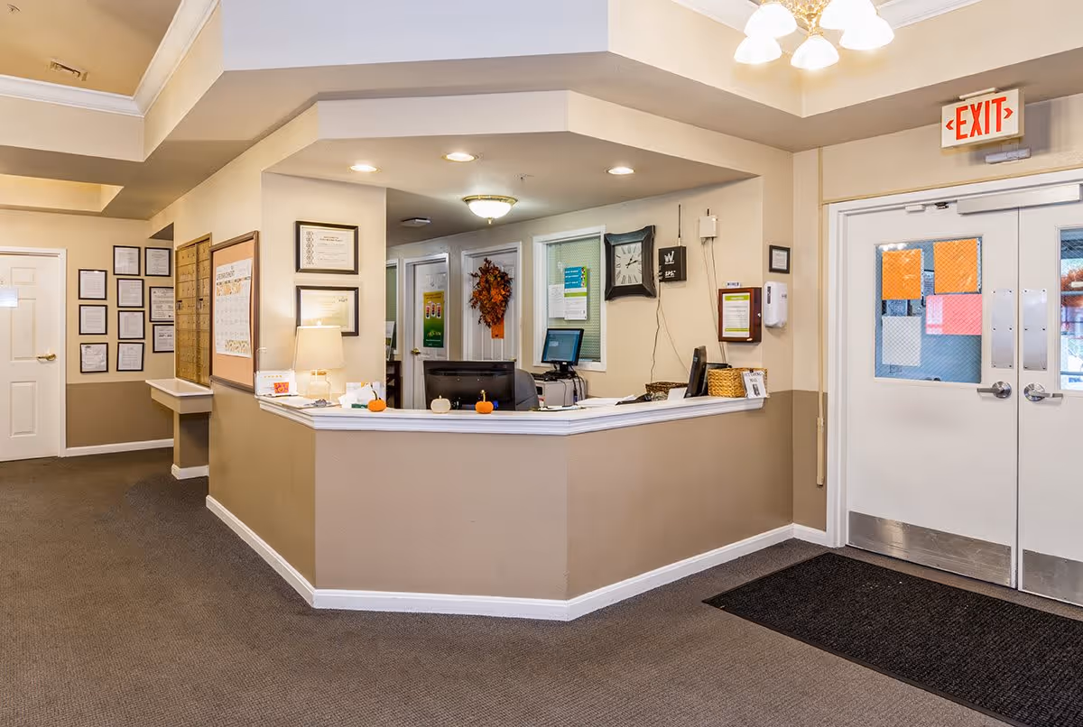 Reception area of The Addison of Clen-Moore Place with a beige and white front desk, computer monitors, a lamp, small decorative pumpkins, framed certificates on the walls, an exit sign above double doors, and a carpeted floor.