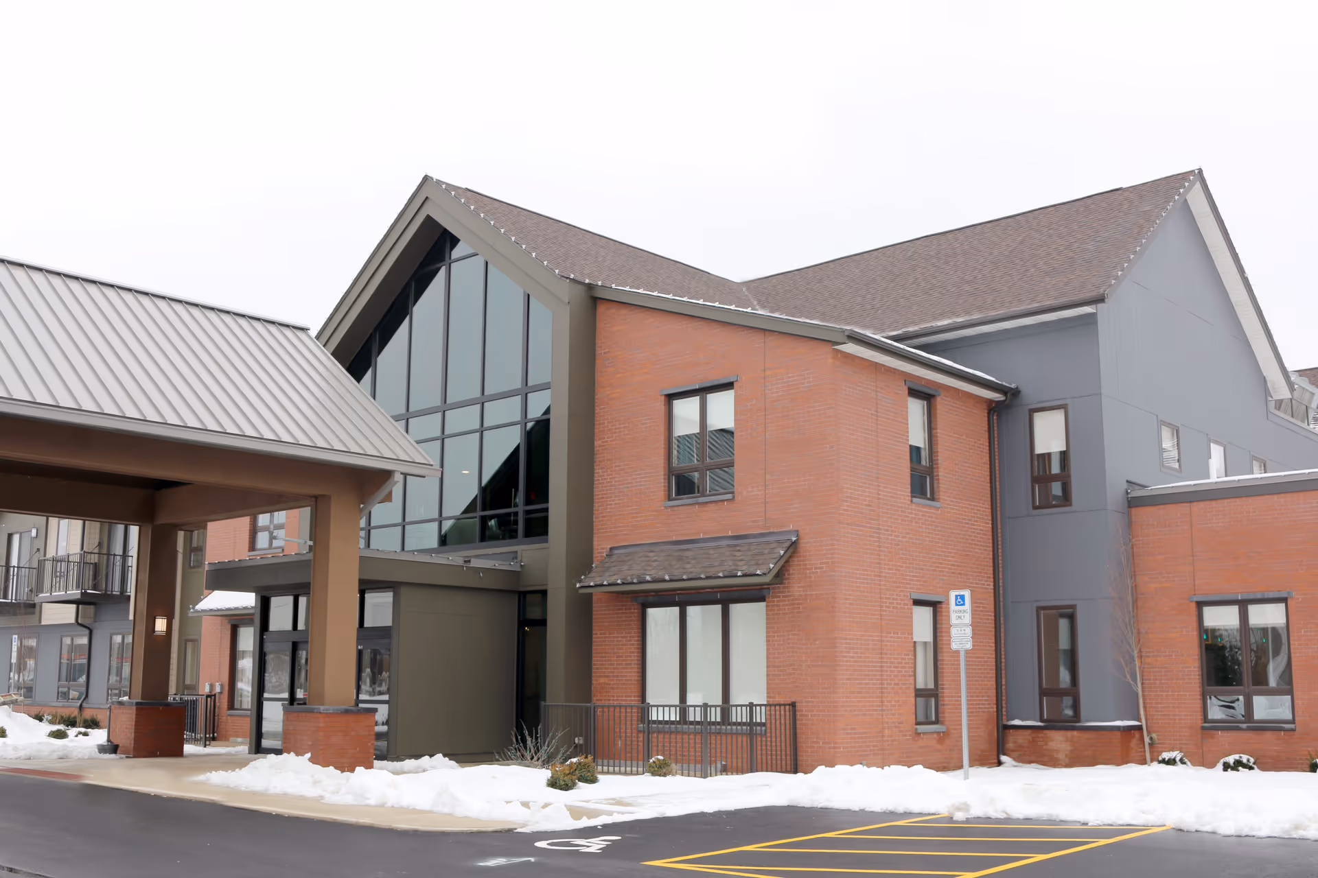 Exterior view of The Patterson senior living facility showing a modern building with large glass windows, brick and gray siding, a covered entrance, and snow on the ground.