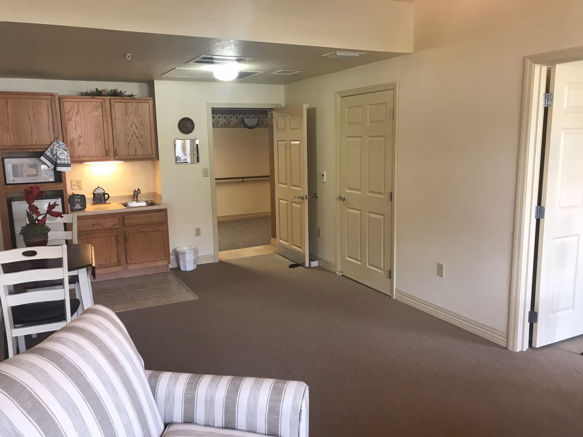 Interior view of a senior living facility room showing a small kitchenette with wooden cabinets, a microwave, a sink, and a countertop. There is a white chair with a red flower arrangement on a table in the foreground, and a striped couch partially visible. The room has beige walls and carpeted floor, with two closed doors and one open door leading to another area.