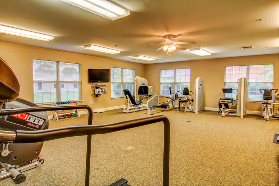 Well-lit fitness room with cardio and strength machines, free weights, and a wall-mounted TV.