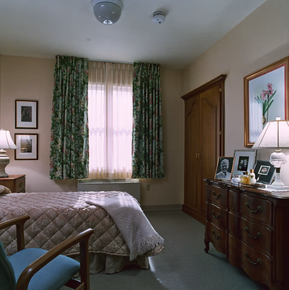 A cozy bedroom with a bed covered in a quilted bedspread and a light blanket. There is a wooden dresser with framed photos, a lamp, and decorative items on top. A wooden wardrobe stands next to the dresser. The window has sheer curtains with floral-patterned drapes. Two framed pictures hang on the wall beside the window. A cushioned chair with wooden arms is partially visible in the foreground.