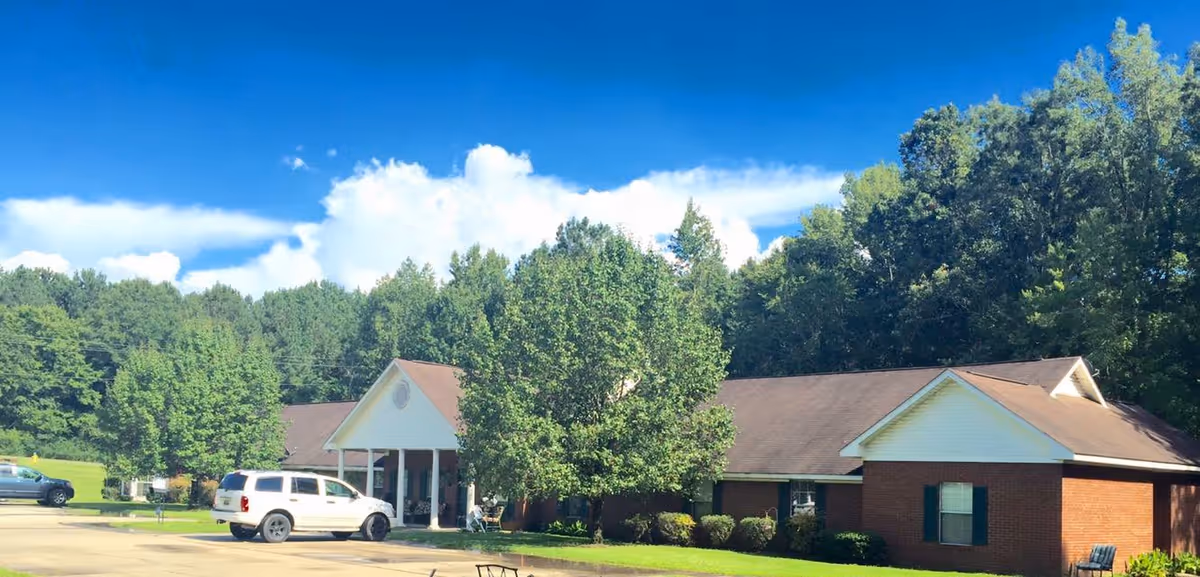 Exterior view of a single-story brick building with a brown roof, surrounded by green trees and a clear blue sky. There are a few parked cars in front of the building and a small lawn area with bushes and trees.