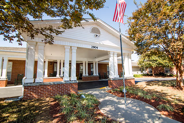 Front entrance of a brick senior living building with a white columned portico displaying the address '2904', an American flag, and landscaped grounds.
