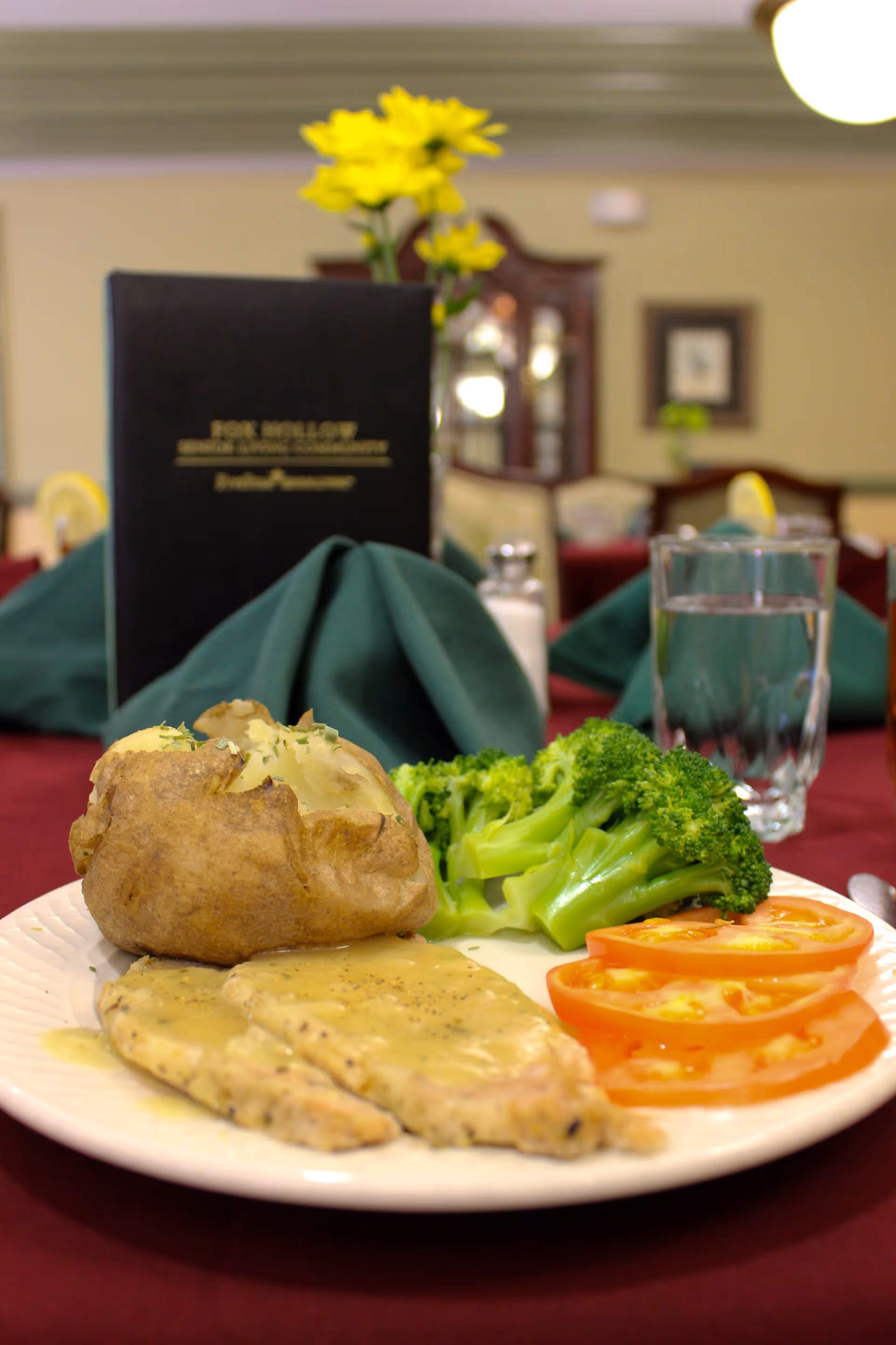 A plated meal on a table with a baked potato, steamed broccoli, sliced tomatoes, and two pieces of chicken with gravy. In the background, there is a glass of water, a salt shaker, a green folded napkin, a menu from Fox Hollow Senior Living Community, and a vase with yellow flowers on a burgundy tablecloth.