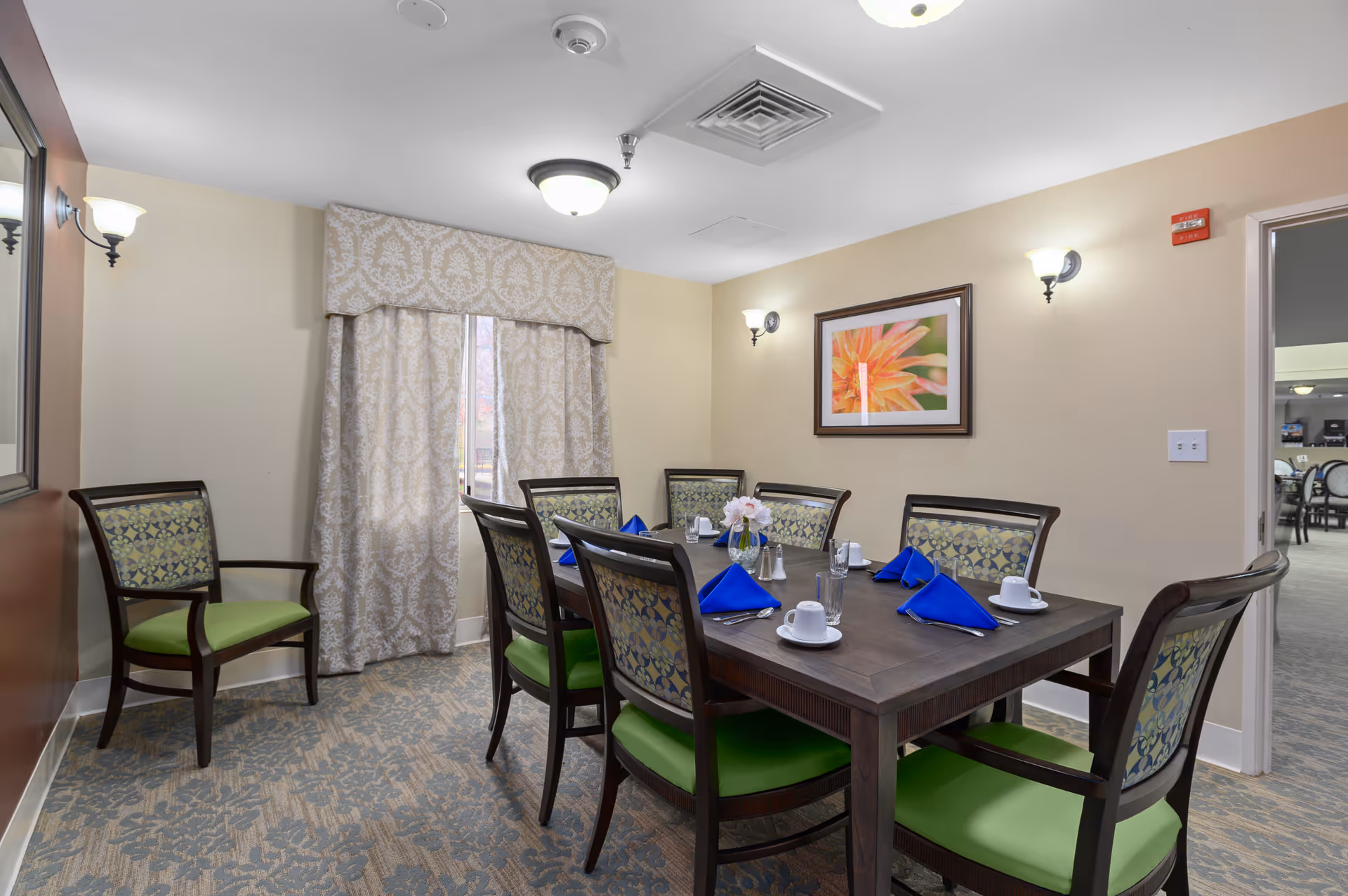 A dining room in a senior living facility with a rectangular wooden table set for six people. The chairs have green cushions and patterned backs. Blue napkins, white cups, glasses, and silverware are neatly arranged on the table. The room has beige walls, a window with patterned curtains, wall sconces, and a framed flower picture on the wall. There is an open doorway leading to another room with more tables and chairs.