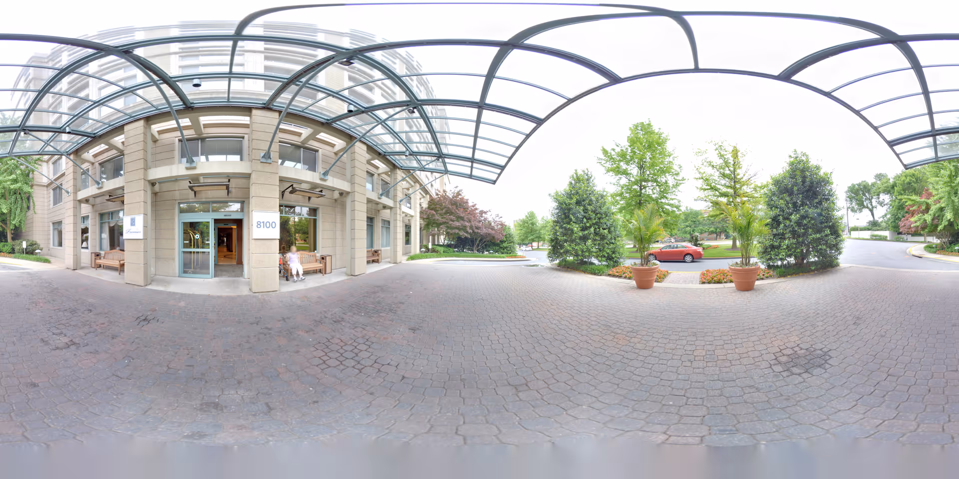 Entrance area of a senior living facility with a covered driveway. The building has large glass doors and windows, with benches on either side of the entrance. There are potted plants and trees around the driveway, and a red car is parked nearby.