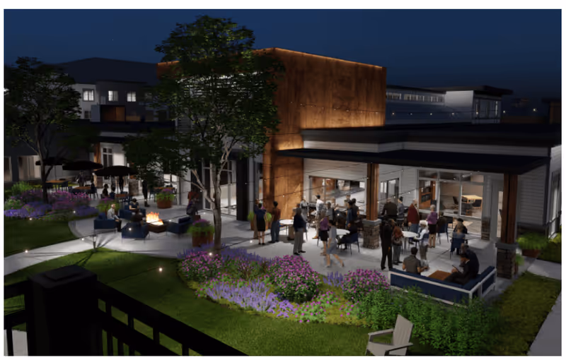 Nighttime exterior view of a modern senior living building with people gathered on a landscaped patio with seating, a fire pit, and outdoor dining areas.