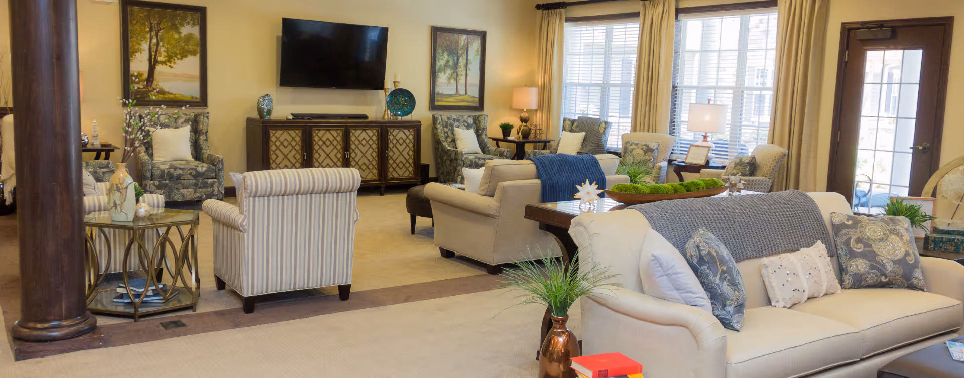 A spacious and well-lit living room area with multiple sofas and armchairs arranged around a central coffee table. The room features large windows with beige curtains, a flat-screen TV mounted on the wall, decorative paintings, and various plants and lamps providing a cozy atmosphere.