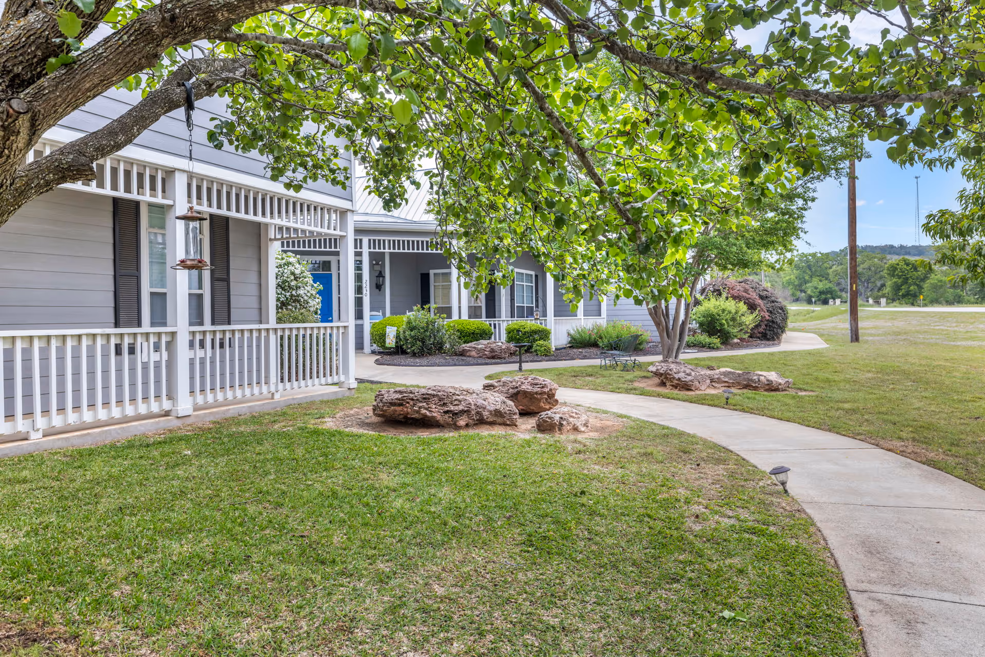 A curved concrete walkway leads through a grassy outdoor area with large rocks and trees, alongside a gray building with white trim and a porch featuring white railings and a blue door.