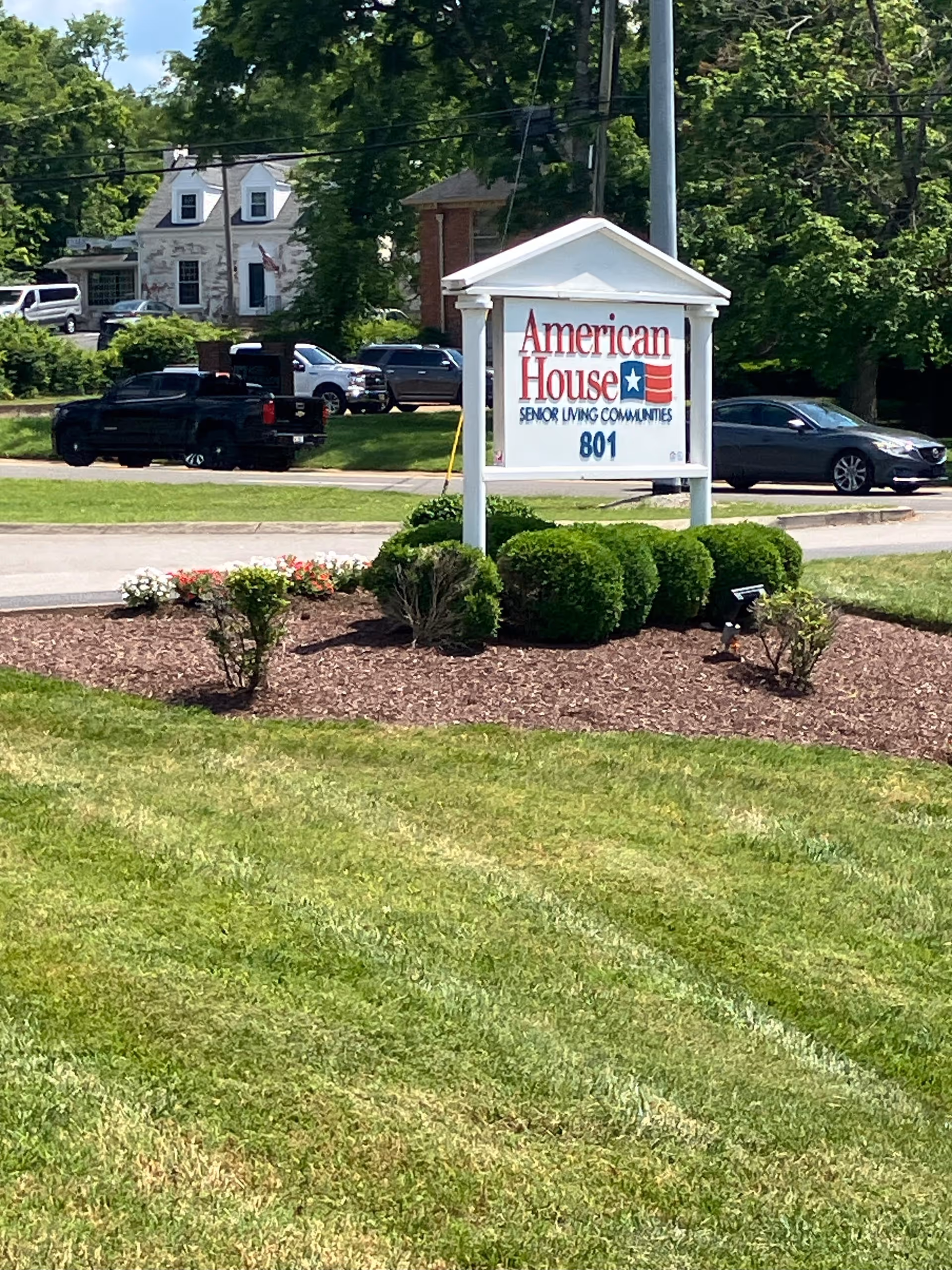 A white roadside sign reading "American House Senior Living Communities 801" on a landscaped lawn with shrubs and parked cars in the background.