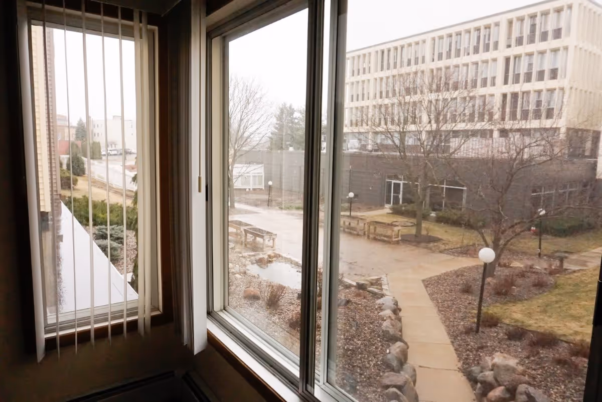 View through a corner window looking out over a courtyard with walkways, benches, lamp posts, and a multi-story building.