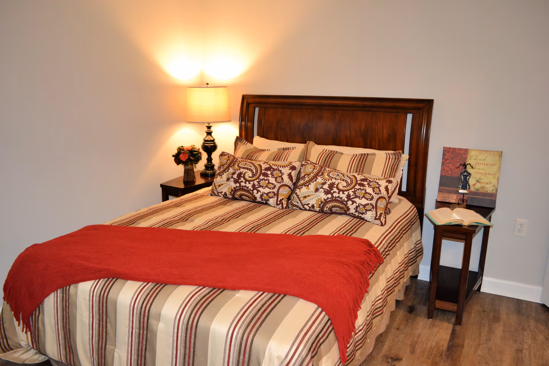Neatly made bed with striped bedding, patterned pillows and a red throw, flanked by a lamp and nightstands in a bedroom.