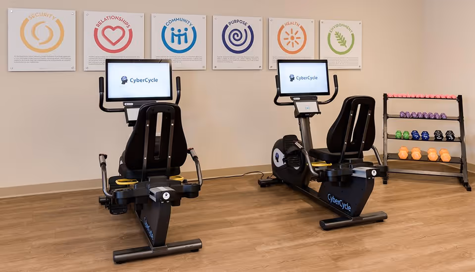 A fitness room with two CyberCycle recumbent exercise bikes facing a wall with six framed posters displaying words and icons related to security, relationships, community, purpose, health, and environment. To the right, there is a rack holding various colorful dumbbells.