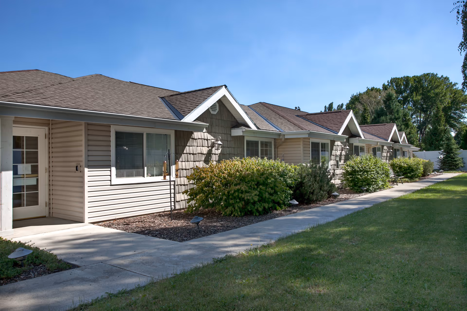 Exterior view of a single-story residential building with beige siding and multiple windows, surrounded by green bushes and a well-maintained lawn under a clear blue sky.