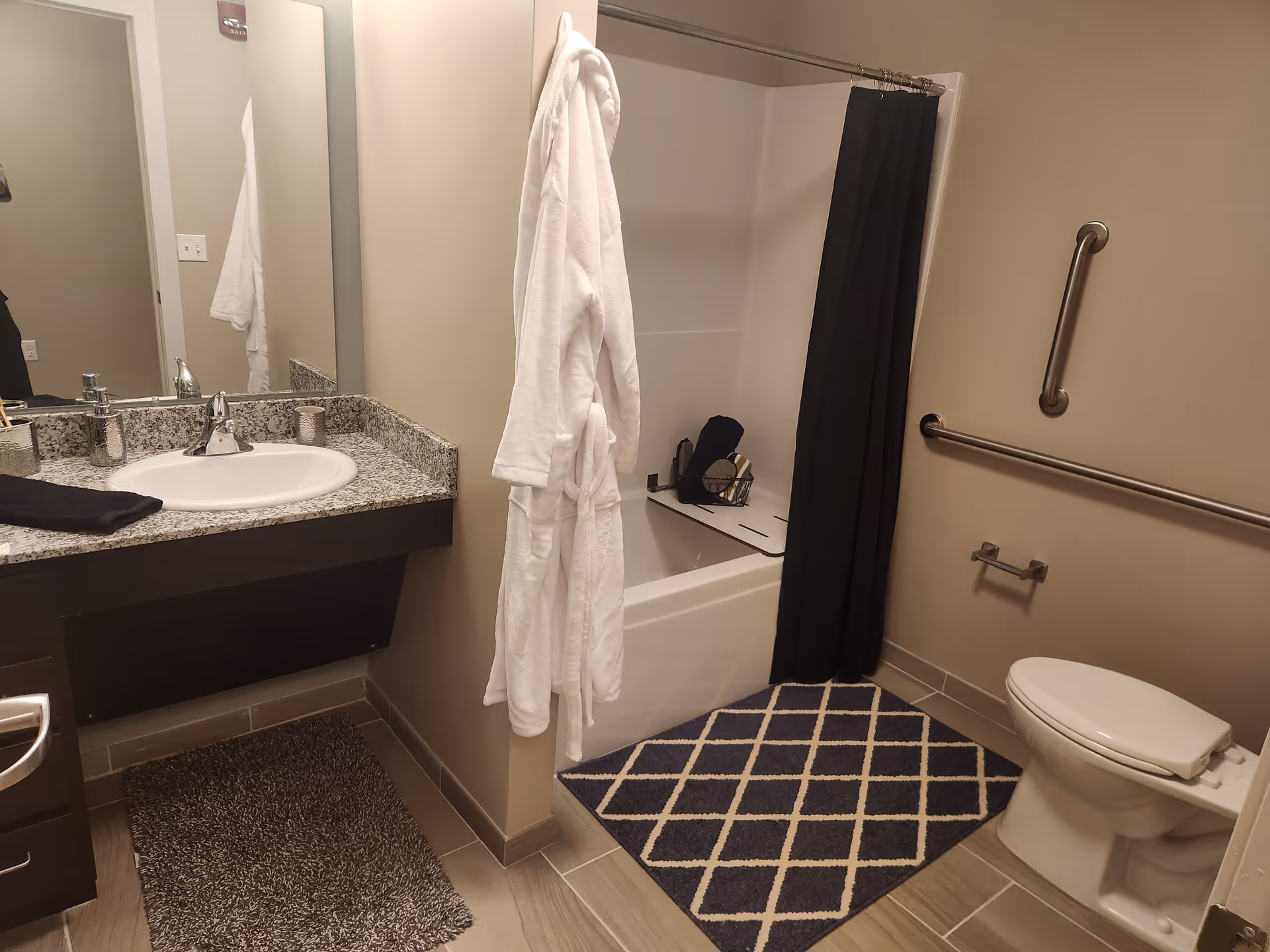 Bathroom with a sink and granite countertop, bathtub/shower with a black curtain, toilet with grab bars, and a white robe hanging on a hook.