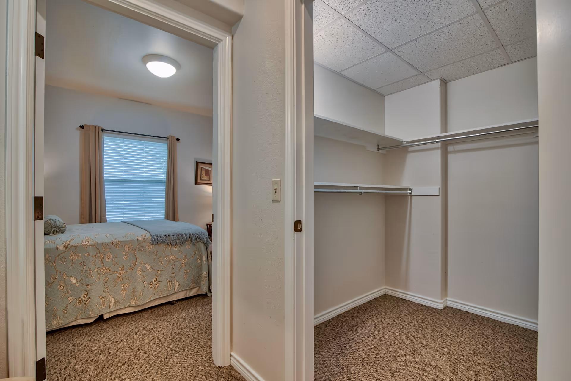 View of a bedroom with a bed covered in a floral bedspread and a window with closed blinds and curtains, seen through an open doorway. Adjacent to the bedroom is an empty walk-in closet with shelves and hanging rods, beige walls, and carpeted floor.