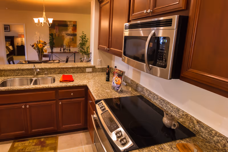 A modern kitchen with granite countertops, wooden cabinets, a stainless steel microwave, and an electric stovetop. There is a double sink on the left side and a cookbook and a bottle of wine on the counter. In the background, a dining area with a chandelier, a table with a flower vase, and a colorful abstract painting on the wall are visible.