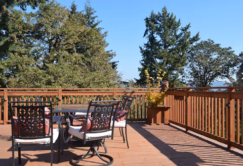 Outdoor wooden deck with a table and six cushioned chairs, surrounded by a wooden railing. Trees and clear blue sky are visible in the background.