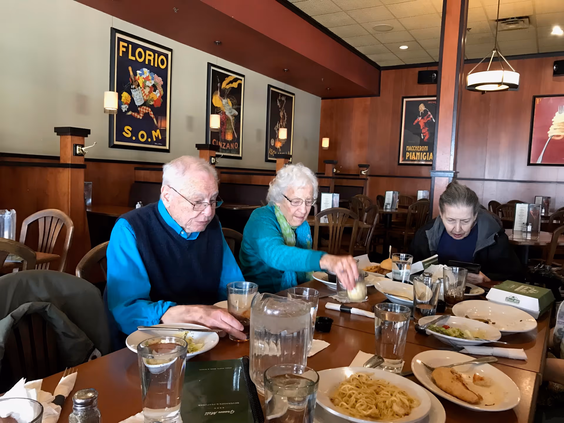 Three elderly people sitting at a wooden dining table in a restaurant, eating and drinking. The table has plates with food, glasses of water, and a pitcher. The background shows wooden panel walls with framed vintage posters and wall-mounted lights.