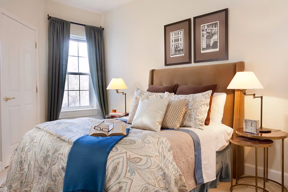 A cozy bedroom with a neatly made bed featuring multiple pillows and a patterned comforter. There is a blue throw blanket at the foot of the bed with an open book and glasses resting on it. Two bedside tables each hold a lamp, and one has a small clock. A window with dark curtains is on the left wall, and two framed black and white pictures hang above the headboard.