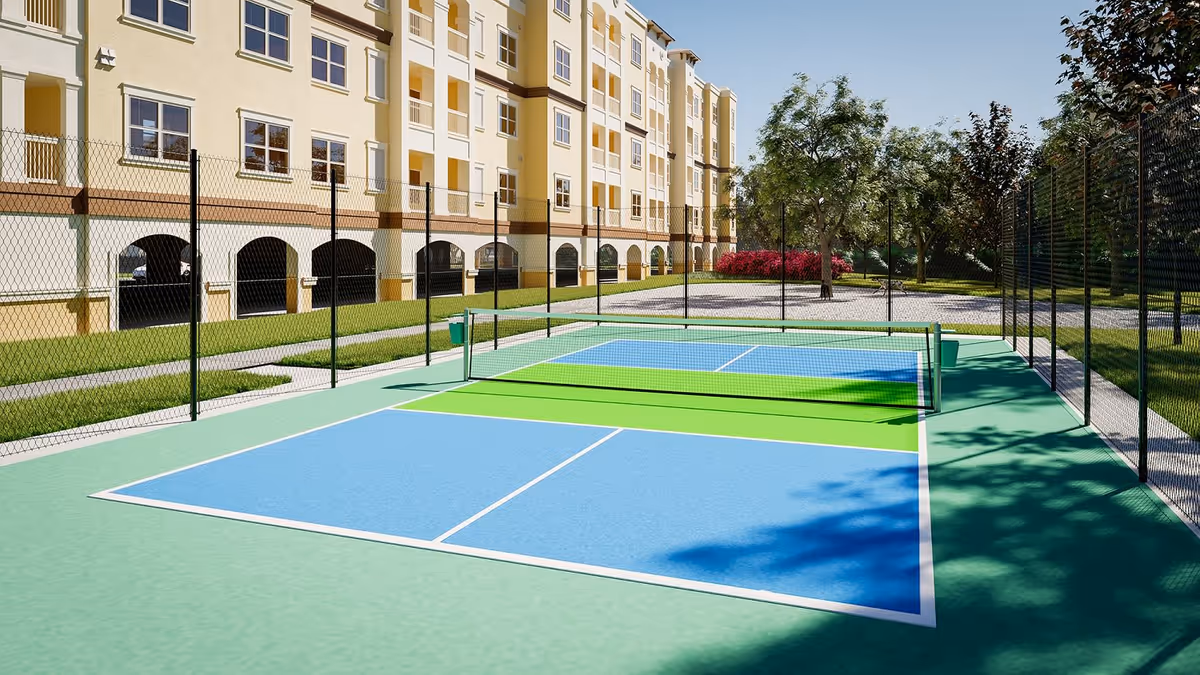A fenced outdoor pickleball/tennis court with blue and green playing surfaces next to a multi-story residential building and landscaped grounds.
