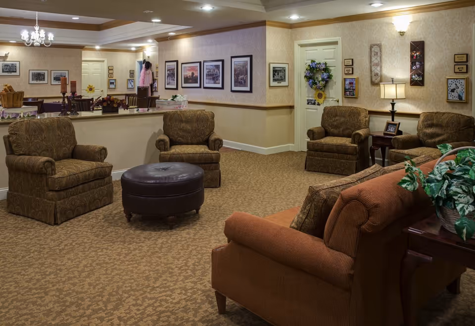 A warm, furnished senior living lounge with armchairs, a sofa, a round ottoman, side tables, wall art and soft lighting.