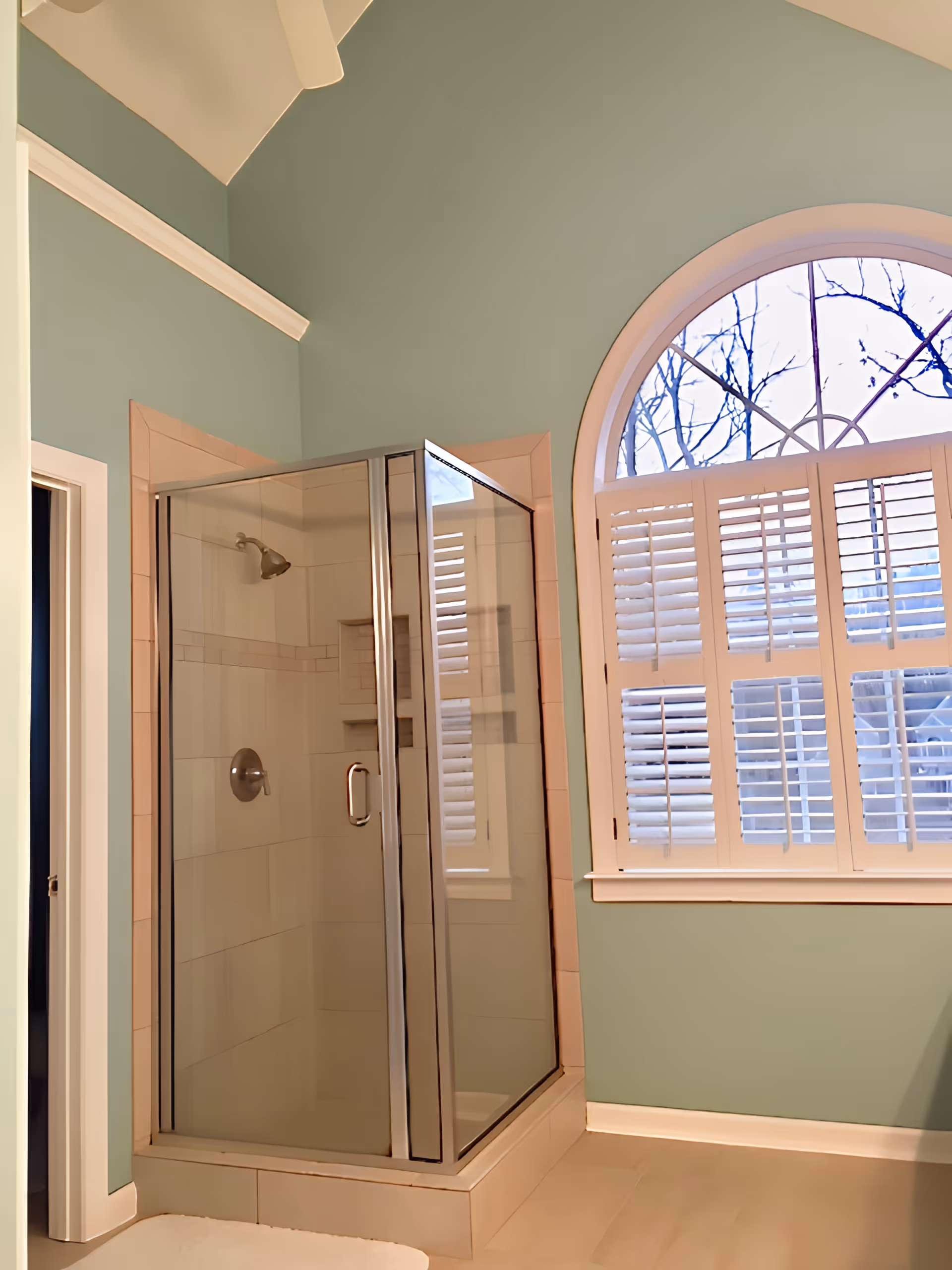 A bathroom with a glass-enclosed corner shower featuring beige tiles and a silver showerhead. The walls are painted light green, and there is a large arched window with white plantation shutters letting in natural light.