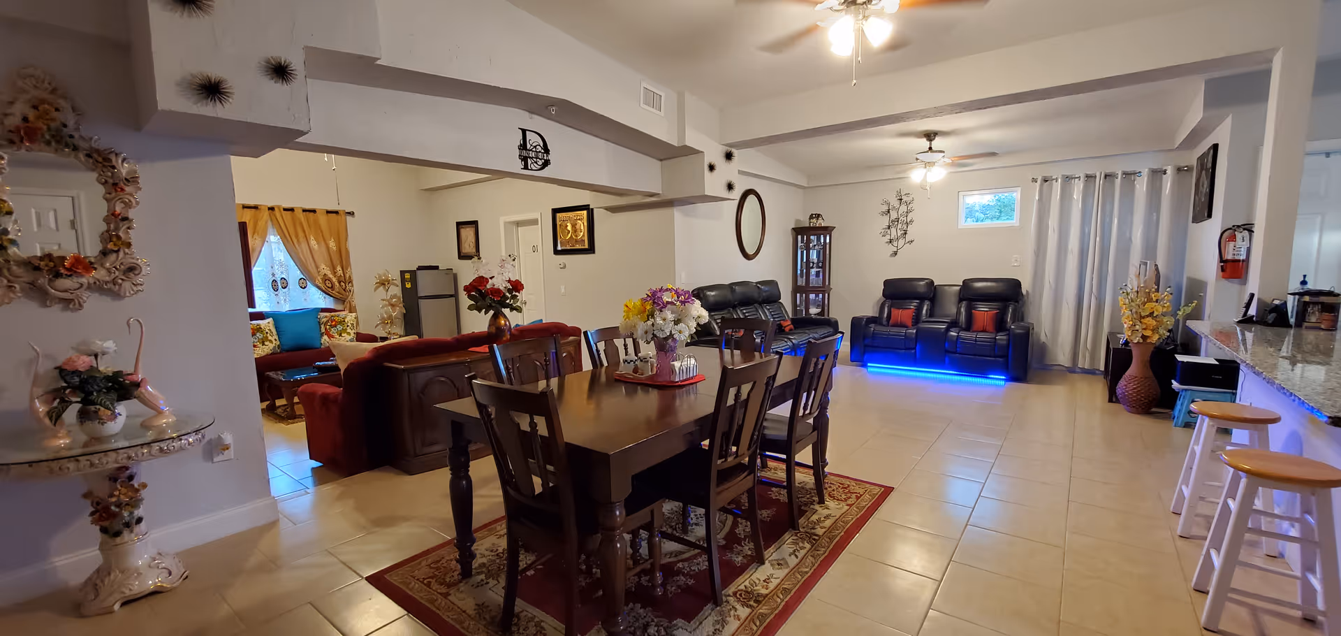 A spacious interior room featuring a dining area with a wooden table and six chairs on a decorative rug. Beyond the dining area, there is a living room with black leather recliners, one with blue LED lighting underneath, and a red sofa set near a window with yellow curtains. The room has tiled floors, ceiling fans with lights, and various decorative items including flowers, wall art, and a mirror. A kitchen counter with bar stools is visible on the right side.