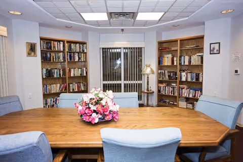 Community reading room with a wooden table surrounded by upholstered chairs, bookshelves and a floral centerpiece.