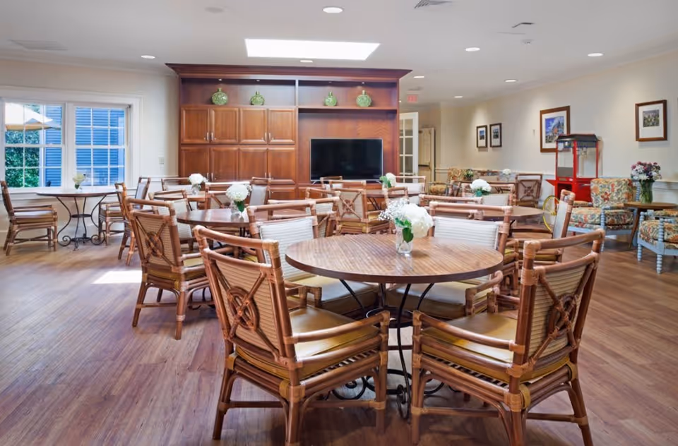 A bright and spacious dining room with multiple round wooden tables surrounded by wicker chairs. Each table has a small vase with white flowers. The room features wooden flooring, a large wooden cabinet with a flat-screen TV, framed pictures on the walls, and a popcorn machine in the corner. Large windows allow natural light to fill the space.