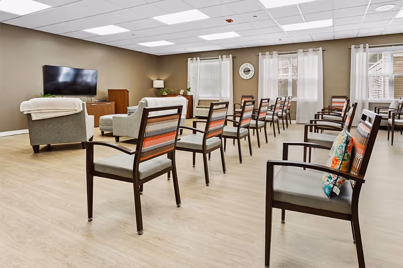 A spacious, well-lit common area with multiple wooden chairs arranged in rows facing a wall-mounted flat-screen TV. The room has light wood flooring, beige walls, and several windows with white curtains letting in natural light. There are also a couple of armchairs and a small cabinet with a lamp and plants.