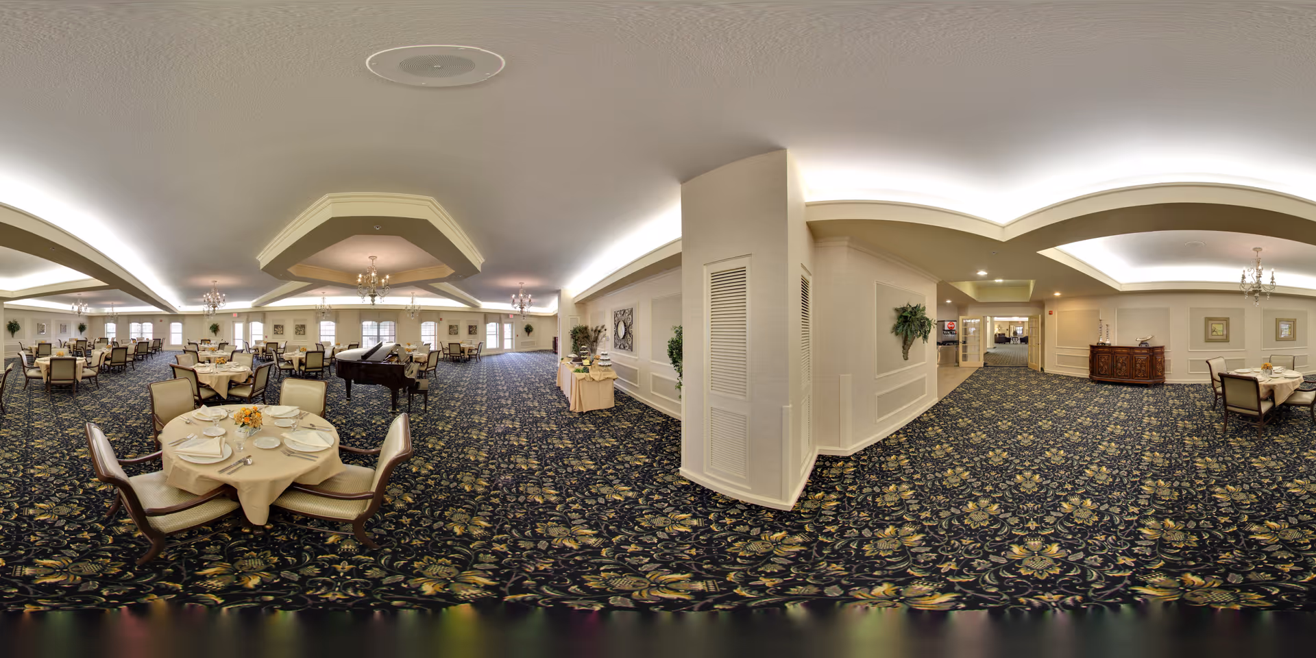A spacious, elegant dining room in a senior living facility with multiple round tables set with white tablecloths, plates, and silverware. The room features a patterned carpet, chandeliers hanging from the ceiling, and a grand piano in the center. The walls are decorated with framed artwork and plants, creating a warm and inviting atmosphere.