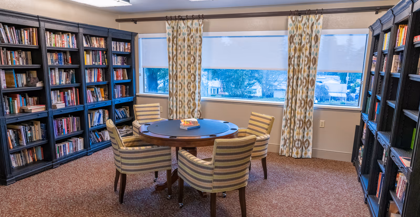 A cozy reading room with dark wooden bookshelves filled with books lining two walls. In the center, there is a round wooden table with a black top and four striped upholstered chairs around it. Two books are placed on the table. Large windows with patterned curtains allow natural light to enter the room.