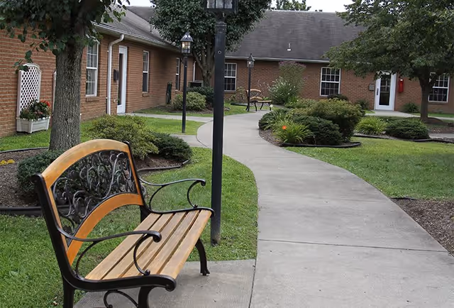 Outdoor courtyard area at Mount Vernon of South Park featuring a curved concrete walkway, a wooden bench with decorative metal armrests, green grass, shrubs, trees, and brick buildings with white doors and windows in the background.