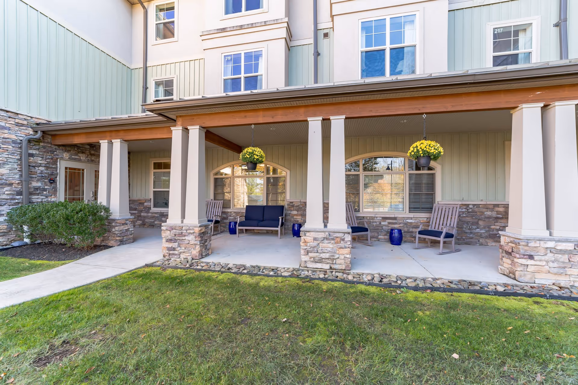 Outdoor covered patio area with stone pillars and wooden beams, featuring several wooden chairs with blue cushions, hanging flower pots with yellow flowers, and a green lawn in the foreground. The building exterior has stone and light green siding with multiple windows.