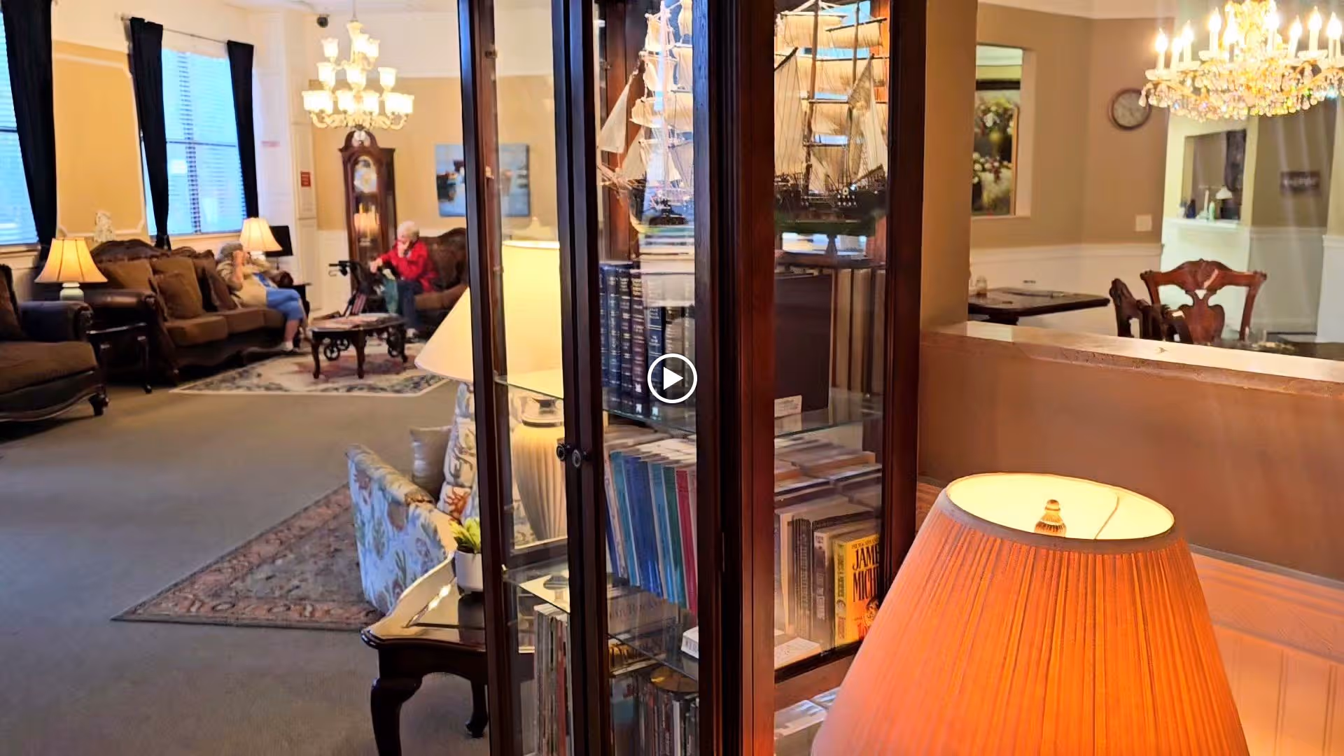 A cozy living room area in an assisted living facility with comfortable sofas, armchairs, a coffee table, and a glass cabinet filled with books and model ships. Two elderly people are seated on the sofas near a window with curtains. The room is warmly lit with table lamps and a chandelier is visible in the adjacent dining area.