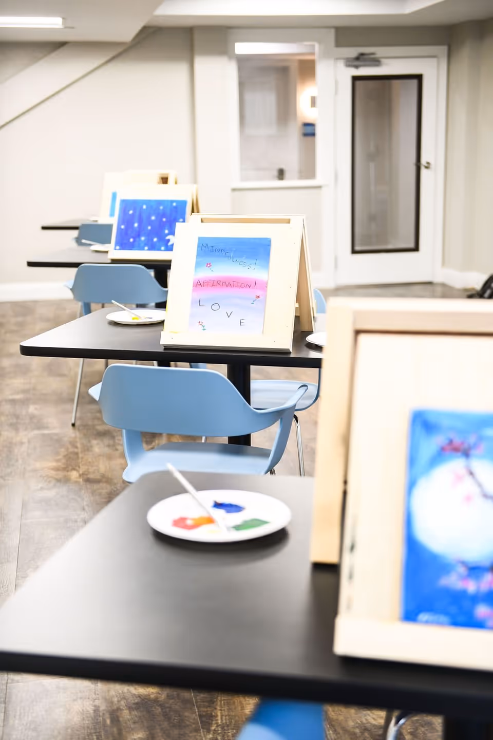 A room with several tables and blue chairs arranged in rows. Each table has a small easel holding a painting or drawing, and a plate with paint and a paintbrush. The paintings include words like 'MINDFULNESS', 'AFFIRMATION', and 'LOVE'. The room has light-colored walls and a door with a window in the background.