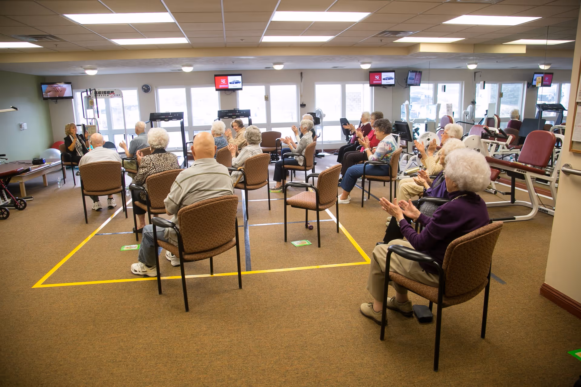 A group of elderly people seated in chairs arranged in a socially distanced manner in a fitness or activity room, participating in a seated exercise or group activity led by an instructor at the front. The room has exercise equipment along the walls and large windows letting in natural light.