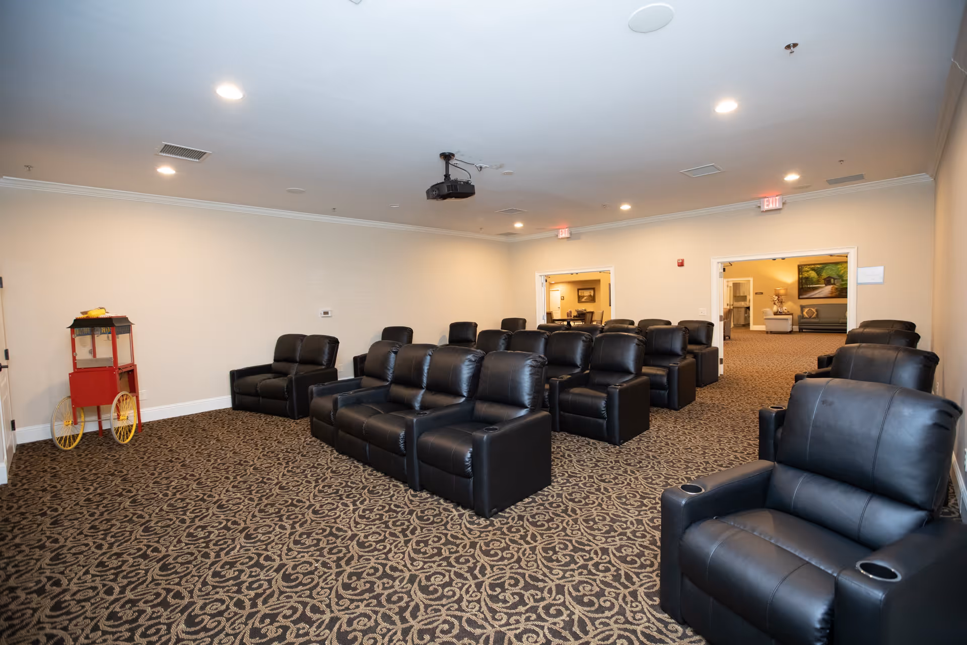 A small theater room with multiple rows of black leather recliner chairs arranged facing a projector mounted on the ceiling. The room has beige walls, patterned carpet, and recessed lighting. There is a red and yellow popcorn machine in the corner and two open doorways leading to other rooms.