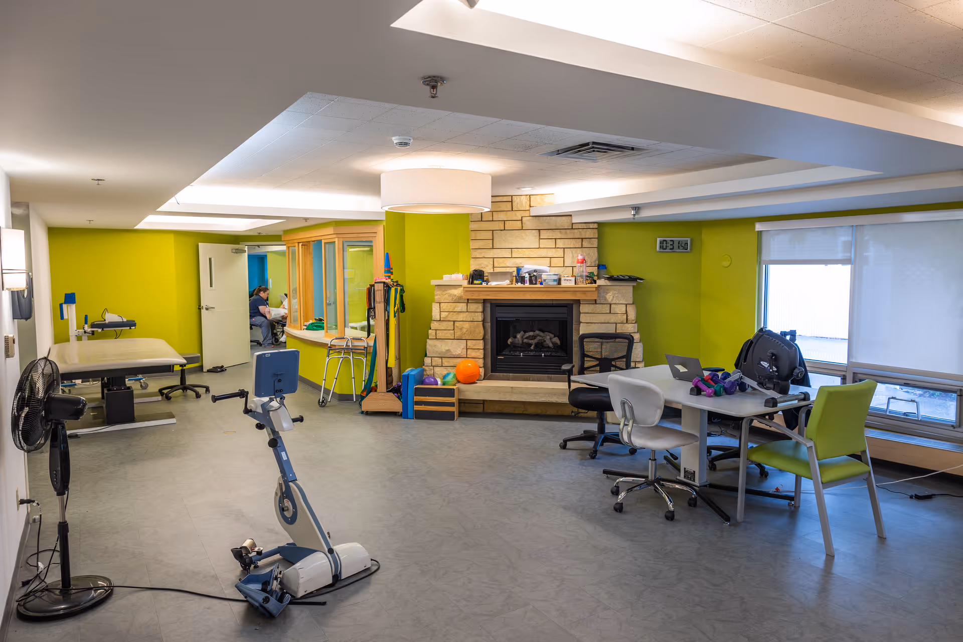 A bright interior activity/therapy room with exercise equipment, a table and chairs, and a stone fireplace against a green wall.