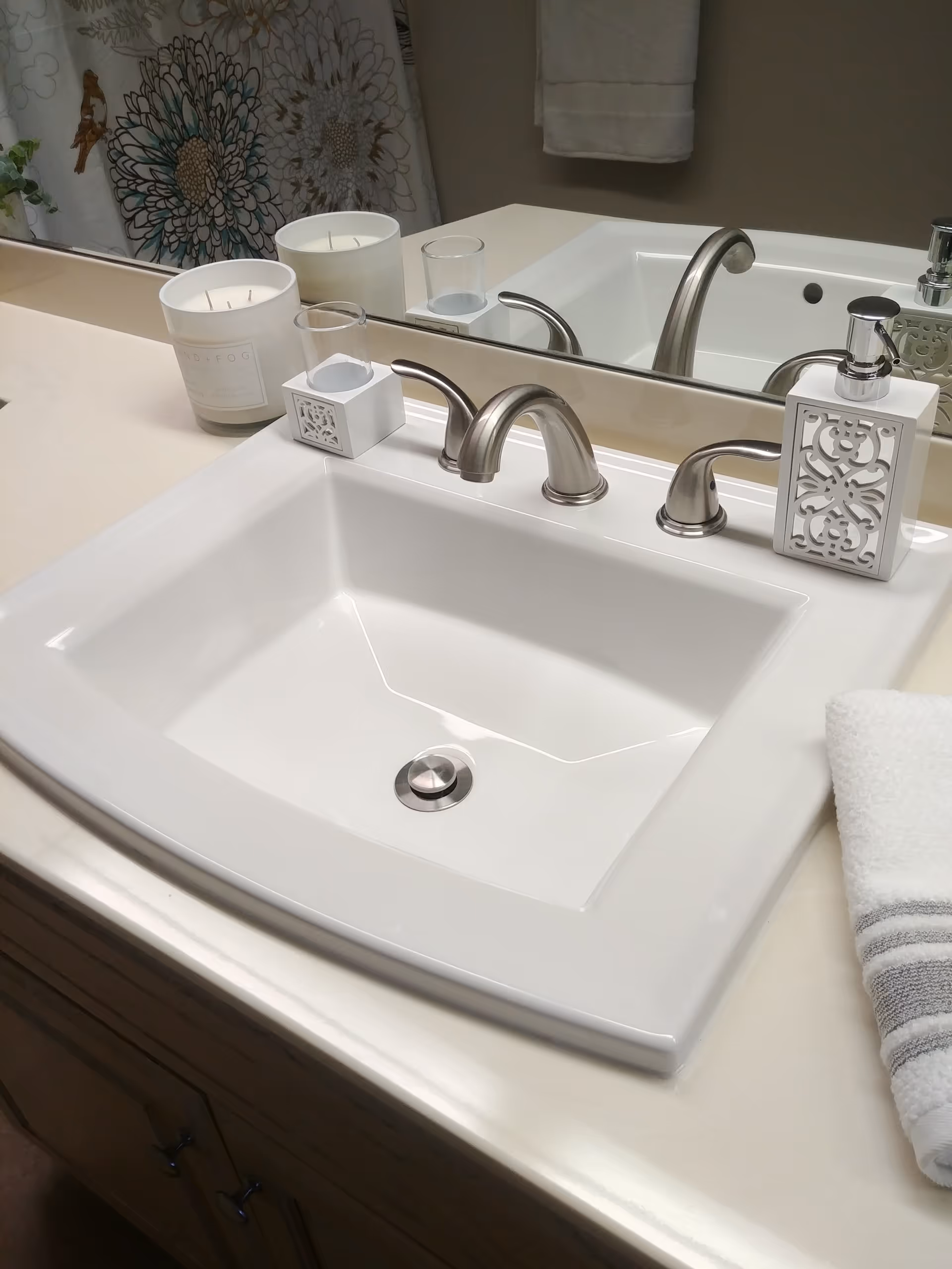 A clean white bathroom sink with a silver faucet, a decorative soap dispenser, a small candle, and a folded white towel with gray stripes on the countertop. A large mirror and a floral shower curtain are visible in the background.