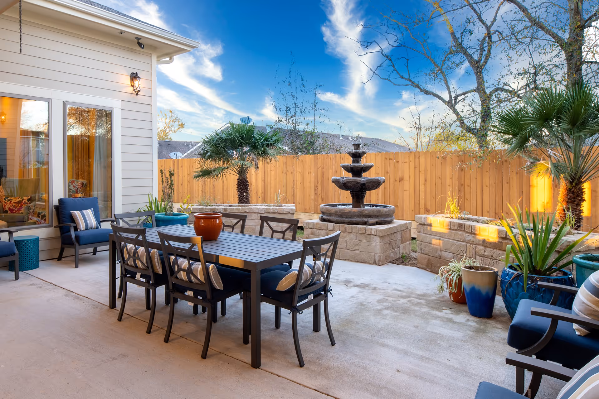 Outdoor patio area with a black metal dining table and six chairs with cushions, surrounded by blue cushioned armchairs and various potted plants. A three-tier stone water fountain is visible against a wooden fence, with trees and a partly cloudy blue sky in the background.